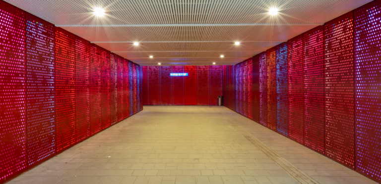 A modern, brightly lit pedestrian tunnel with vivid red perforated walls and a grid-patterned ceiling. The floor is paved with light-colored tiles. Small ceiling lights illuminate the tunnel, creating reflections on the walls.