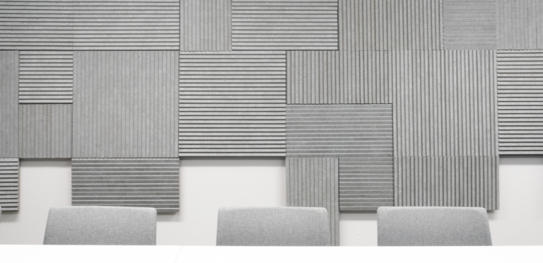 Three gray chairs are aligned in front of a wall adorned with textured, geometric acoustic panels in varying shades of gray, creating a modern and minimalist aesthetic.