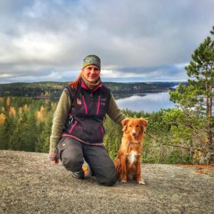 Henkilö, jolla on pääpanta, polvistuu ruskeanvalkoisen koiran viereen kalliolla. Niiden takana on luonnonkaunis näkymä metsäiseen maisemaan, jossa on järvi ja pilvinen taivas.