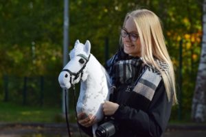 Pitkät vaaleat hiukset ja lasit omaava henkilö hymyilee valkoiselle harrastushevoselle. Heillä on yllään musta takki ja raidallinen huivi. Kamera roikkuu heidän kaulassaan. Taustalla on vihreitä puita ja aita.