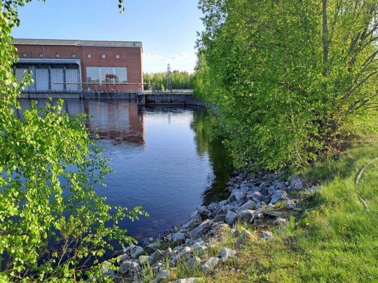 Näkymä rauhalliselle joelle, jota reunustavat vasemmalla tiilirakennus ja oikealla vehreät puut. Etualalla on kivinen nurmialue kirkkaan sinisen taivaan alla.