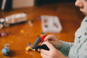 Lapsi vaaleansinisessä paidassa kokoamassa värikkäitä LEGO palikoita puupöydälle. Sumealla taustalla näkyy lisää LEGO-osia ja osittain rakennettu malli.